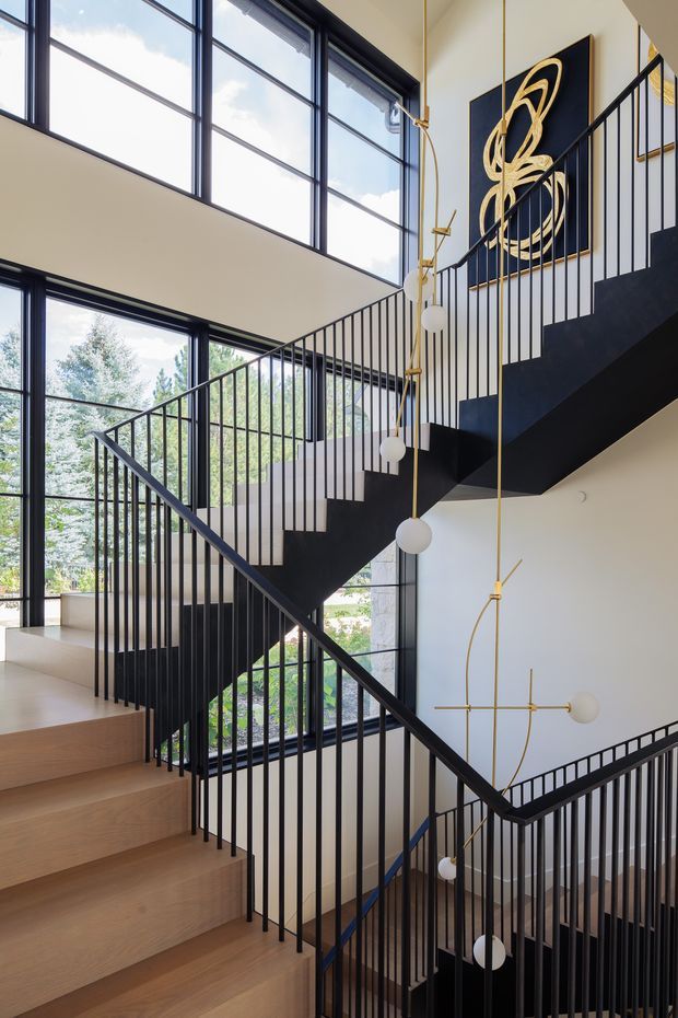 A contemporary multi-level stairway with light timber treads, slim black metal balustrades, expansive black-framed windows, and sculptural brass-and-white globe pendant lighting descending through the stairwell. White walls and abundant glazing create a bright, open vertical space, with abstract black-and-gold artwork visible on the upper landing.