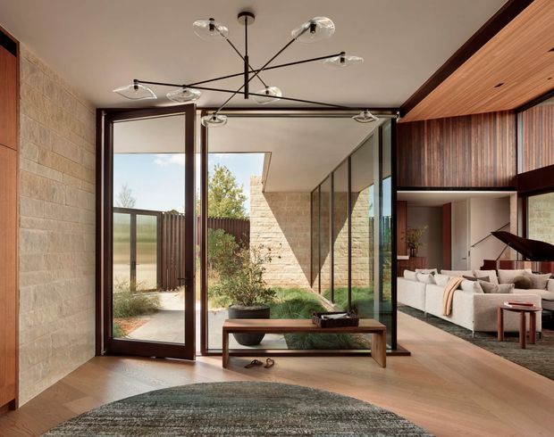 A refined contemporary entryway opens through full-height glazed doors into a landscaped courtyard and an adjoining living room. The interior features warm timber flooring, natural stone block walls, dark timber-framed glazing and cabinetry, a timber slatted ceiling, built-in wood-lined joinery, a sculptural black chandelier, and a neutral upholstered sofa. A bench and textured rug define the foreground entry space, while the courtyard brings in abundant natural light.
