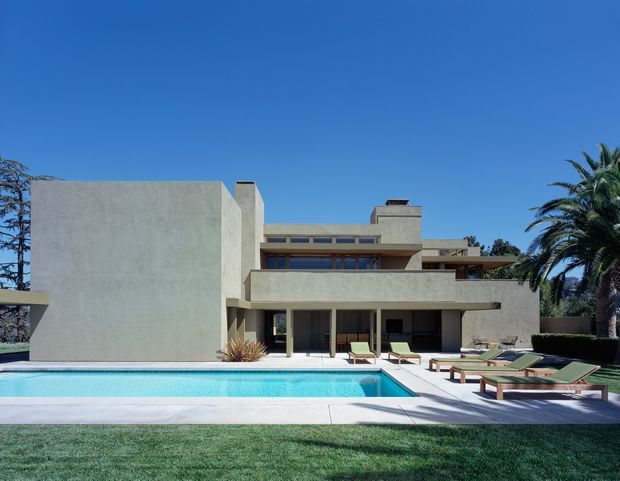 Exterior view of a large modernist flat-roofed residence with smooth pale stucco walls, strong horizontal and vertical geometric volumes, deep overhangs, and extensive glazing. A rectangular swimming pool and paved terrace with loungers occupy the foreground, with manicured lawn and mature palm trees surrounding the property under a clear blue sky.