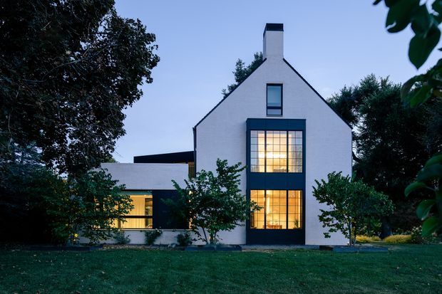 Front exterior view of a contemporary two-storey house at dusk, framed by mature trees and lawn. The home has a simple steep gable roof, white rendered walls, a dark chimney, and tall black-framed glazed openings glowing warmly from within. A lower flat-roofed wing extends to the left.