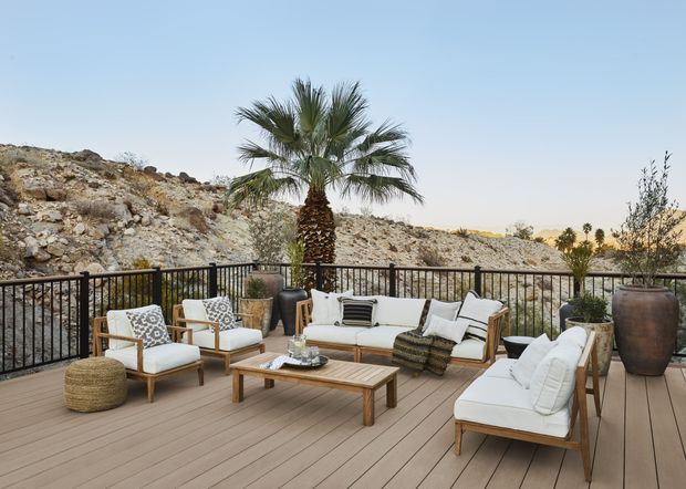 A spacious outdoor terrace furnished as a relaxed lounge, with a timber-look deck, white upholstered sofas and armchairs, a low wooden coffee table, woven ottoman, decorative planters, and a black metal balustrade. A palm tree and rugged rocky terrain are visible beyond the railing under a clear sky, creating a warm desert or arid landscape setting.