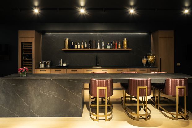 A sophisticated dark-toned home bar and entertaining kitchen with a large stone island, integrated timber cabinetry, a backlit dark feature wall, open bottle shelving, wine storage, and three upholstered bar stools with brass-toned frames. Recessed spotlights create a dramatic, luxurious atmosphere.