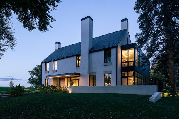 Twilight exterior view of a substantial multi-storey home set within a landscaped garden. The building has pale rendered walls, a steep dark pitched roof with multiple tall chimney forms, large vertically proportioned windows, and a projecting glazed corner volume glowing warmly from interior lighting. A low rendered boundary wall and steps define the foreground terrace or garden edge, while mature trees frame the property.