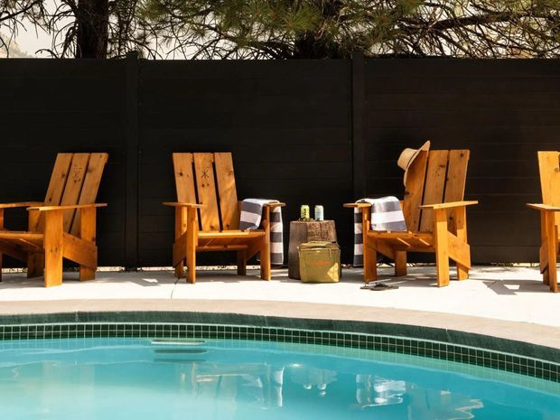 Outdoor poolside scene with a curved turquoise swimming pool in the foreground, pale paved pool deck, and several timber Adirondack-style lounge chairs arranged against a dark privacy fence. Small side tables and outdoor accessories add a relaxed residential leisure setting, with trees visible above the fence.