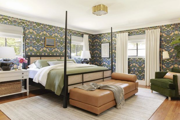 A bright, traditionally styled bedroom with a four-poster bed, floral botanical patterned wallpaper, pale curtains, and light wood flooring. The room includes a large upholstered ottoman at the foot of the bed, a green armchair, white bedside furniture, framed artwork, and a brass-toned ceiling light. Multiple windows provide abundant natural light.