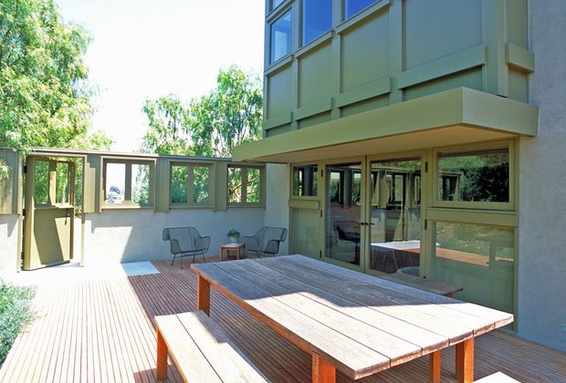 A sunlit elevated timber deck courtyard beside a modern multi-level home. The space features a large built-in timber dining table with benches, additional outdoor lounge chairs, extensive green-framed glazing and doors, and a projecting upper-storey volume clad in pale green panels. Mature trees surround the outdoor area.