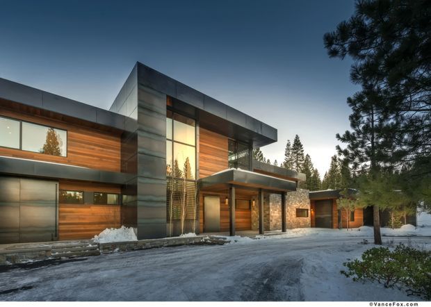 A contemporary multi-level residence in a snowy, forested setting. The building combines horizontal warm timber cladding, dark metal roof planes and framing, large glazed openings, and a prominent vertical stone-clad entry volume. A covered entry and driveway are visible in the foreground, with pine trees and snow surrounding the property.
