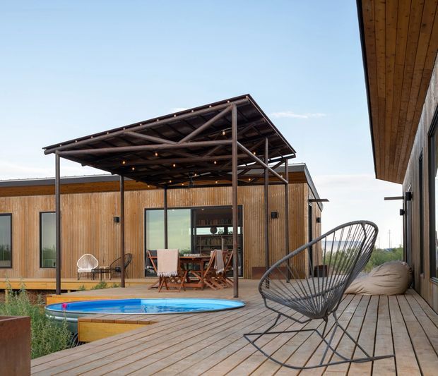 Outdoor residential entertaining area with a timber-clad single-storey building, expansive timber decking, a covered pergola, outdoor dining furniture, and a small round swimming pool. The scene has a relaxed contemporary pavilion-like design with exposed dark metal pergola framing and warm timber surfaces.