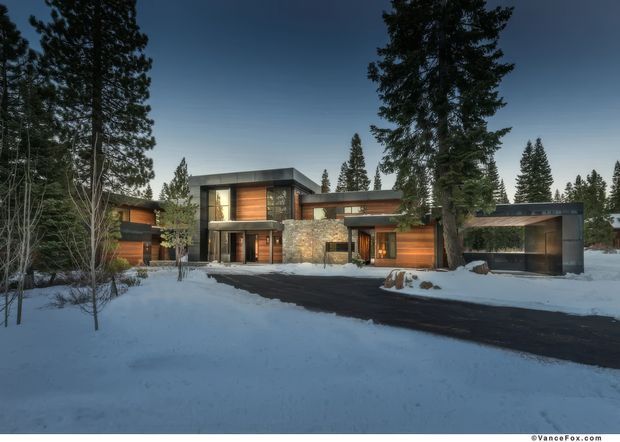 Exterior view of a contemporary multi-level home set in a snowy, wooded landscape. The building features dark horizontal cladding, warm timber garage and wall panels, extensive glazing, natural stone masonry, strong horizontal rooflines, and a detached-looking dark garage or carport structure connected to the main residence.