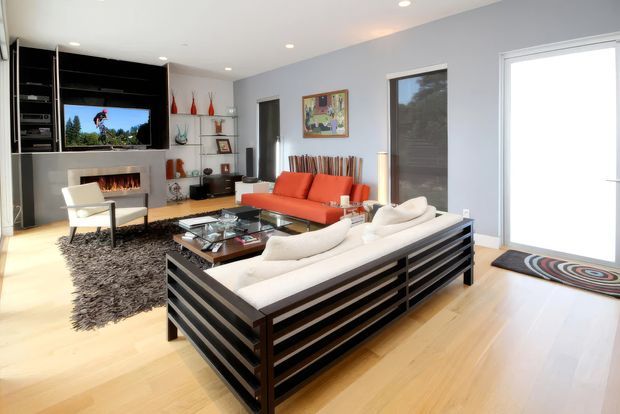 Bright contemporary living room with pale timber flooring, light grey walls, a black built-in media and fireplace wall, and two contrasting sofas arranged around a glass coffee table. Large windows and a glazed door provide abundant natural light, while colourful artwork, sculptural accessories, and a patterned rug add visual accents.