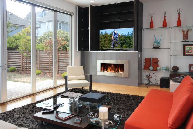 Contemporary living room with floor-to-ceiling sliding glass doors opening to a small garden, a built-in grey media wall with television and linear fireplace, open display shelving, and a vivid orange-red sofa. A glass coffee table and dark textured rug complete the polished, high-contrast interior.