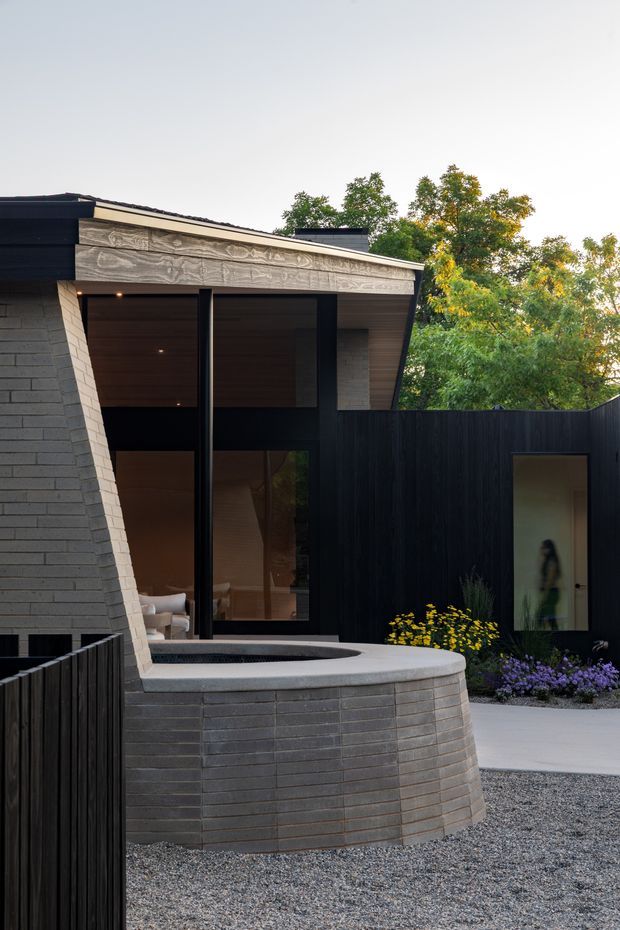 Exterior architectural photograph of a contemporary residential courtyard. The composition features dark grey brick walls, a dramatic angled roofline with a weathered timber fascia, tall black-framed glazing, black vertical timber fencing, and a curved built-in brick water feature or raised planter in the foreground. Gravel paving and planted garden beds surround the building, while warm interior lighting and furnishings are visible through the glazing.