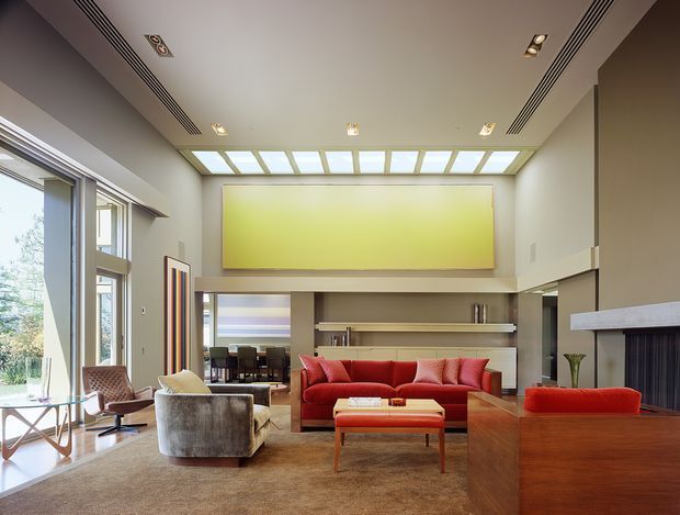 A spacious, refined living room with a high ceiling and clerestory-style roof glazing above a large abstract feature panel. The room features floor-to-ceiling windows, a red sectional sofa, sculptural armchairs, a timber coffee table, built-in shelving, and a substantial fireplace with a pale stone surround. The palette combines warm timber and carpet tones with neutral walls and vivid red upholstery.
