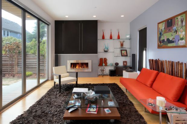 Modern living room with a bright orange-red sofa, white armchair, dark shag rug, glass-topped coffee table, built-in dark cabinetry, open shelving, and a wall-mounted fireplace. Full-height sliding glass doors open toward a small outdoor area, while colorful artwork and sculptural decor add visual accents.