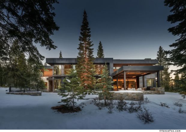 Exterior view of a modern multi-level home set in a snowy, forested landscape. The building features strong horizontal lines, flat roof sections, extensive floor-to-ceiling glazing, dark metal or painted frames, warm timber cladding, and a covered outdoor entertaining area supported by stone-clad elements. Evergreen trees and snow surround the house.