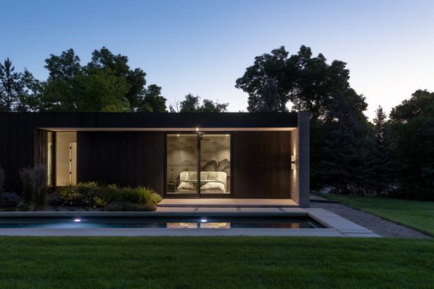 A modern single-storey pavilion-style house at dusk, set within a landscaped garden. The dark timber-clad facade has a low flat roof, broad floor-to-ceiling glazing, and a bedroom visible through the central window. A narrow reflecting or swimming pool runs along the front, bordered by paving and lawn.