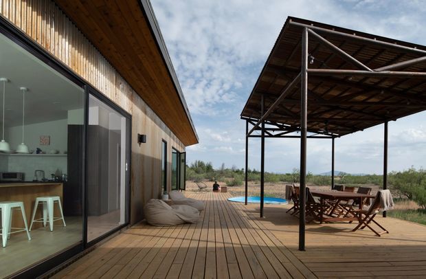 A contemporary single-storey timber-clad house opens onto a broad timber deck with a covered outdoor dining and lounge structure. Large sliding glass doors reveal a compact kitchen with a timber island and stools. The deck overlooks a natural, sparsely vegetated landscape and includes a glimpse of a blue swimming pool.