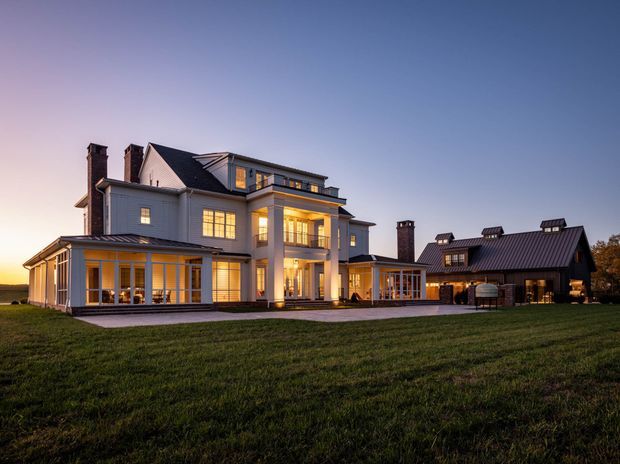Exterior architectural photograph of a large multi-storey residence at dusk. The main house has a light painted facade, dark standing-seam pitched roofs, multiple chimneys, extensive illuminated glazing, covered verandah-like areas, and a substantial connected dark-roofed wing. It sits within a broad lawn, with warm interior lighting creating a welcoming evening atmosphere.