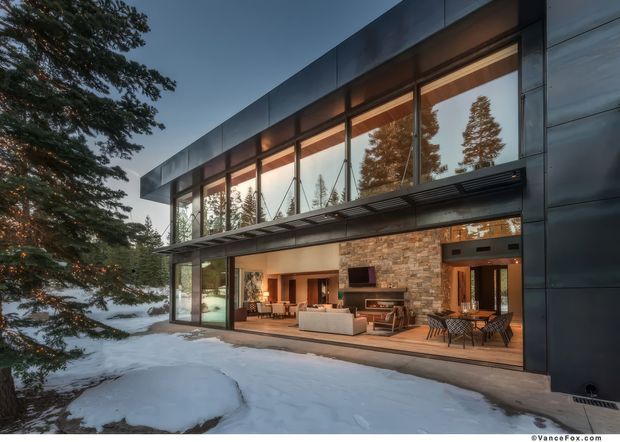 Exterior view of a contemporary two-storey home set in a snowy, wooded landscape. The building features a dark metal-clad facade, extensive floor-to-ceiling glazing, exposed warm timber soffits, and a large open ground-floor living area with a stone feature wall, kitchen, dining area, and outdoor-style seating visible through the glazing. Snow-covered ground and mature evergreen trees surround the house.