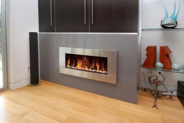 Contemporary living room feature wall with a long built-in fireplace set into a dark grey panel, framed by dark wood cabinetry with slim vertical handles. Pale timber flooring, a tall black speaker, and open glass shelving displaying decorative objects complete the space.