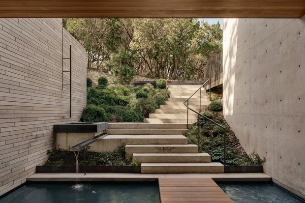 A refined courtyard-like outdoor space viewed beneath a timber-lined overhead structure, with a narrow reflective pool crossed by a timber footbridge in the foreground. Broad concrete steps rise through layered landscaping between a pale horizontal brick wall on the left and a smooth exposed concrete wall on the right, creating a calm, integrated indoor-outdoor composition.