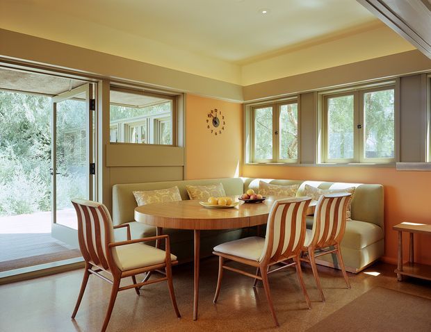 Warm, mid-century-inspired dining and sitting area with a round timber dining table, upholstered striped dining chairs, and a built-in or low-profile sofa beneath a bay of timber-framed windows. The room has peach-beige walls, a recessed tray ceiling with downlights, polished warm-toned flooring, and generous glazing opening toward a leafy exterior.