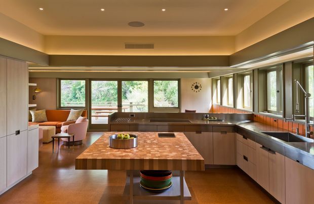 Warm, spacious open-plan kitchen with a large timber butcher-block island, extensive wood cabinetry, stainless-steel benchtops, and abundant windows. A connected lounge area with seating is visible beyond the kitchen, while recessed lighting and a low, broad ceiling create an inviting mid-century-inspired atmosphere.