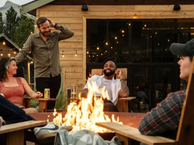 A warm outdoor gathering beside a timber-clad building, with several people seated around a large open fire table. String lights glow across the patio, and a wide dark-framed glazed opening reveals the interior behind them. The scene has a relaxed, social outdoor-lounge atmosphere with mountain terrain visible in the background.