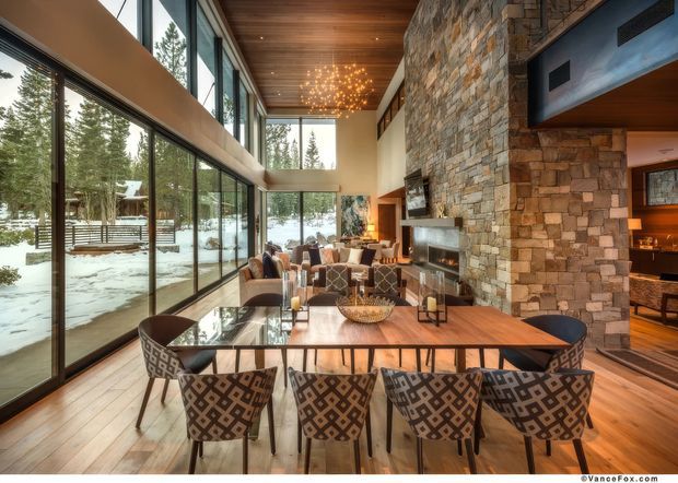 A spacious open-plan dining and living area with a long timber dining table and patterned upholstered chairs in the foreground. Floor-to-ceiling glazing overlooks a snowy forested landscape, while a large natural stone-clad fireplace wall, timber ceiling, warm timber flooring, and contemporary furnishings create a refined alpine lodge atmosphere. A kitchen or bar area is visible beyond the stone feature wall.