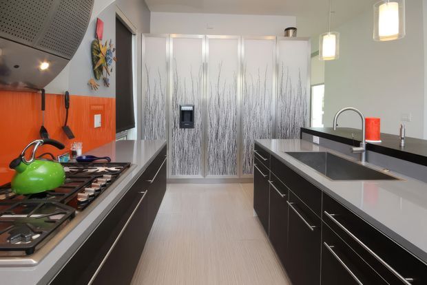 A contemporary galley-style kitchen with dark glossy cabinetry, pale grey engineered-stone benchtops, a large island with an inset sink and curved chrome tap, and integrated appliances. The rear cabinetry features a distinctive frosted botanical-patterned finish, while the cooking wall has a vivid orange glass splashback and stainless-steel rangehood. Pendant lights and a light-toned floor brighten the compact, streamlined space.