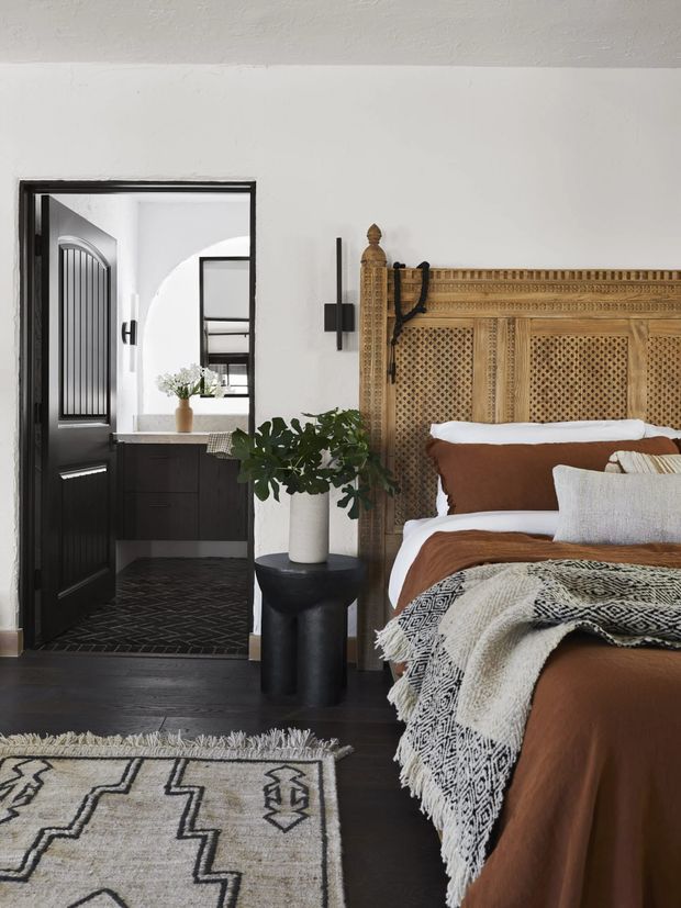 A warmly styled bedroom with a large ornate woven wood headboard, brown bedding, layered patterned throws, and a geometric area rug over dark timber flooring. A black-framed doorway opens directly to an ensuite bathroom with dark cabinetry, a pale stone benchtop, black patterned floor tiles, a rounded wall arch, and a black-framed arched-panel door. White walls and ceiling provide contrast, while a potted plant and black pedestal table add natural and sculptural accents.