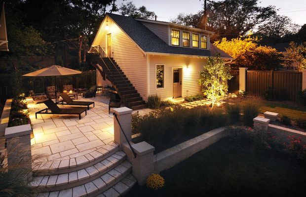 Twilight exterior view of a compact multi-level house with a steep gable roof, dormer windows, light horizontal weatherboard cladding, and warm interior lighting. A paved terrace with outdoor seating, umbrella, steps, retaining walls, landscaped garden beds, fencing, and illuminated planting surround the home.