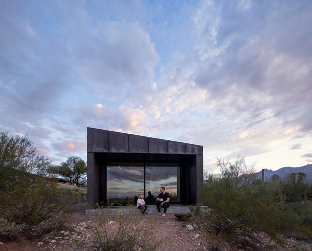 A compact dark-clad pavilion set within a rugged desert landscape, photographed at dusk. The rectilinear structure has a deep overhanging roof or parapet, dark metal-like exterior panels, and a large floor-to-ceiling glazed opening reflecting the sky and landscape. Two people sit on the entrance threshold. Sparse desert vegetation, rocky ground, distant mountains, and a dramatic cloud-filled sky surround the building.