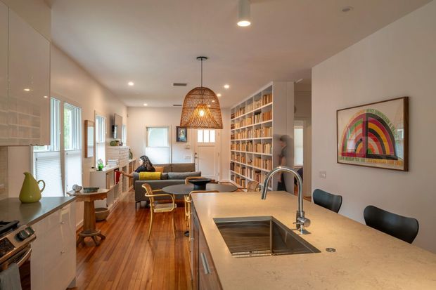 Open-plan kitchen and living area in a long, light-filled interior. The kitchen features pale stone benchtops, white built-in cabinetry, a large undermount sink with gooseneck tap, and dark seating. Beyond the island is a warm timber-floored lounge with a sofa, lounge chairs, and a full-height built-in bookcase. A woven pendant light hangs over the island, while colourful framed artwork adds a playful accent. Windows and glazed doors bring natural light into the space.