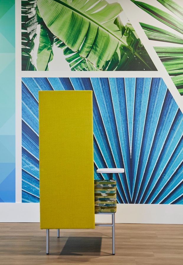 A vibrant contemporary interior vignette featuring a tall lime-yellow upholstered lounge chair with slim metal legs, positioned in front of oversized botanical and abstract blue-green graphic wall artworks. A small striped side table sits beside the chair. The room has a light timber floor, white framing and trim, and a bright, playful high-contrast palette.