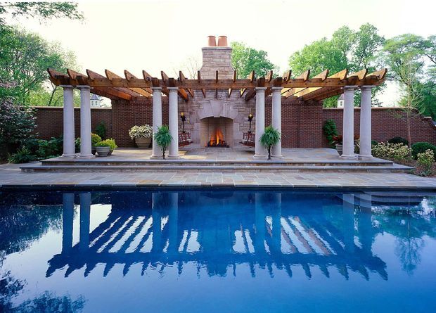 Formal outdoor pool terrace with a classical brick and stone fireplace pavilion. A timber pergola with exposed rafters is supported by evenly spaced white columns, while a paved terrace fronts a deep blue swimming pool that reflects the structure. Brick boundary walls, clipped planting, and a lit open fireplace create a refined traditional garden setting.