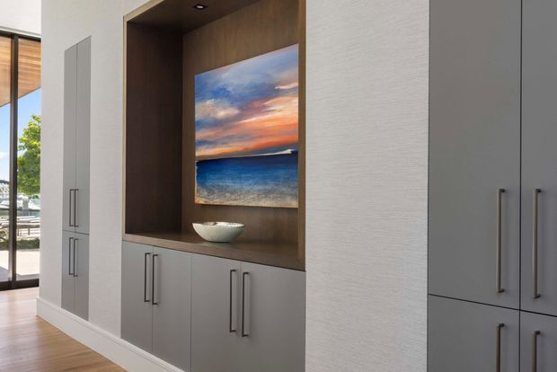A modern built-in storage and display wall in a bright interior, featuring matte grey cabinetry with long dark handles, a warm timber-lined recessed niche, and a coastal landscape artwork above a small bowl. Floor-to-ceiling glazing at left brings in daylight and reveals an outdoor setting. The cabinetry has clean, minimalist lines and integrated joinery.