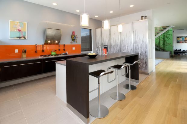 Bright contemporary kitchen with a large dark island and integrated breakfast bar, four modern metal stools, glossy dark cabinetry, and a vivid orange glass backsplash. The room combines pale tiled flooring in the kitchen zone with warm timber flooring extending into the adjacent living area. White pendant lights, recessed ceiling lights, and a decorative grey branch-like feature wall add visual interest, with a glimpse of a green-accented adjoining space beyond.