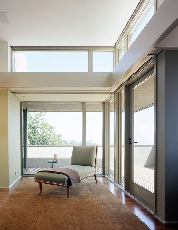 A bright, refined sitting area with a mid-century-style lounge chair and matching ottoman positioned on a large neutral rug. Full-height glazed doors and clerestory windows open toward a terrace or balcony, creating abundant natural light. The room has pale walls, warm timber flooring, slim dark window frames, and clean architectural lines.