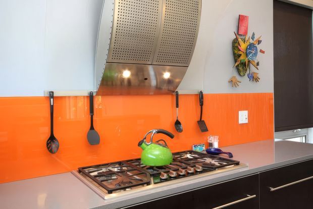 Bright contemporary kitchen viewed close-up, featuring a large gas cooktop with a green kettle, dark lower cabinetry, pale benchtops, and a vivid glossy orange tiled splashback. A stainless steel perforated rangehood with integrated lighting dominates the upper area, with cooking utensils hanging against the splashback and a colourful wall artwork at right.