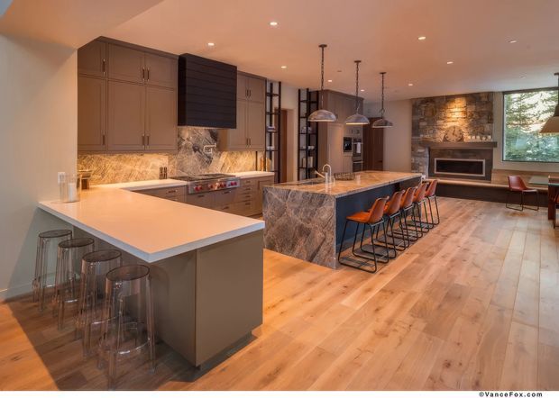 Spacious open-plan kitchen with two large islands, extensive dark cabinetry, marble-look stone benchtops and splashback, pendant lighting, timber flooring, and a dining and living area beyond. A stone-clad fireplace wall and large window are visible in the adjoining living space.