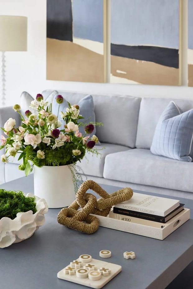 A softly styled contemporary living room vignette featuring a pale grey upholstered sofa with quilted cushions, abstract neutral artwork, a dark coffee table, a large floral arrangement, stacked design books, a woven sculptural object, and a small decorative board game. The palette is light, muted, and refined with natural and tactile accents.