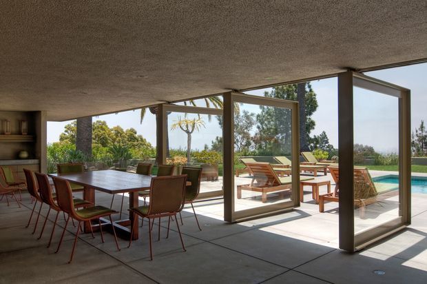 A mid-century-inspired open-plan dining and outdoor entertaining area with a large rectangular dining table and vintage-style chairs under a low textured ceiling. Full-height glazed sliding or folding doors open directly to a paved terrace with timber outdoor seating and a visible swimming pool. The space has strong indoor-outdoor connection, exposed concrete-style flooring, dark structural window frames, and abundant daylight.