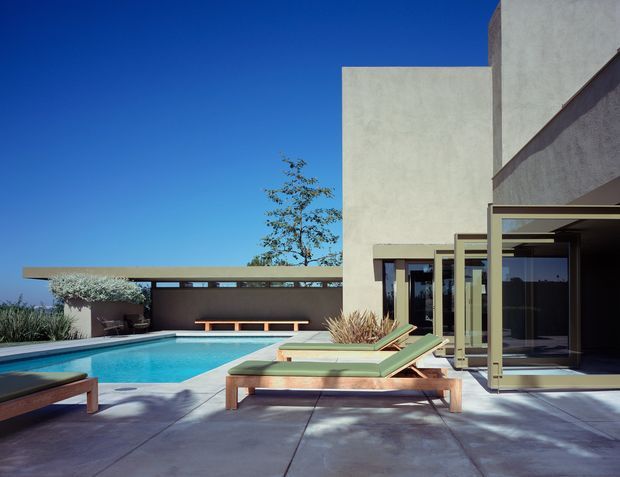 A modern flat-roofed concrete home opening onto a sunlit pool terrace. The composition features expansive glazing and open framed doors, pale rendered walls, broad concrete paving, turquoise swimming pool water, and streamlined timber-framed outdoor loungers with green cushions. Low planting and a deep blue sky reinforce the clean, resort-like outdoor setting.
