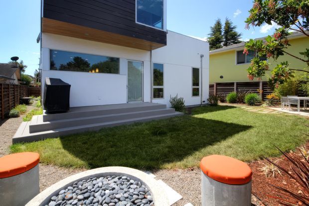Contemporary two-storey residential exterior viewed from a landscaped backyard. The white facade has large windows and a glazed door, a projecting upper level clad in dark horizontal boards with a timber soffit, and broad concrete steps leading to the lawn. A circular pebble garden feature with cylindrical orange-topped seats occupies the foreground, with a neighbouring colourful dwelling visible at right.