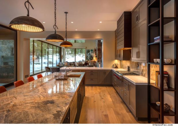 A spacious, warm contemporary kitchen with a large polished stone island, dark wood cabinetry, open shelving, and pendant lighting. The kitchen opens to a dining and living area through extensive glazing, with timber flooring and a forested exterior visible beyond.