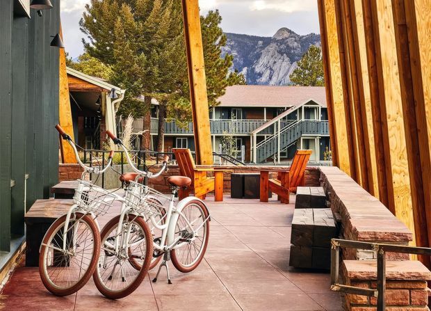 A rustic outdoor courtyard or terrace framed by timber-clad buildings, with several white bicycles in the foreground, wooden Adirondack-style chairs and a table, and built-in timber-and-stone seating along the edge. A mountain landscape is visible beyond the surrounding lodge-style buildings. The space has warm natural timber, dark green cladding, paved flooring, and a relaxed alpine retreat atmosphere.