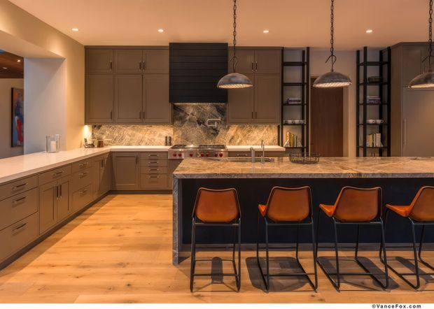 Warm, spacious contemporary kitchen with extensive dark wood cabinetry, a large central island with a stone benchtop, integrated seating for four, stainless steel appliances, open shelving, and pendant lights. The kitchen features timber flooring and a dramatic stone backsplash extending behind the range.