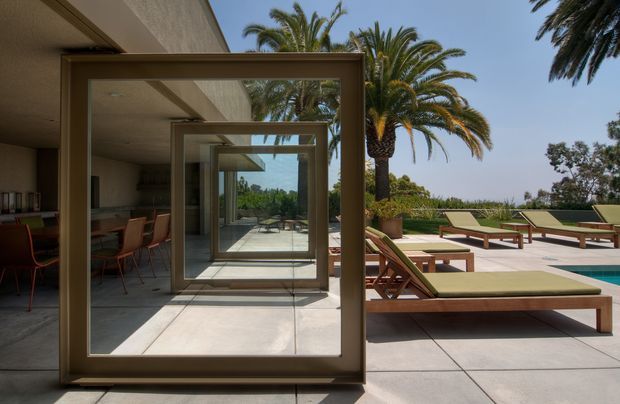 A refined modern outdoor terrace beside a pool, viewed through a sequence of large bronze-toned framed glazed doors. The covered dining area opens onto a pale paved sun terrace furnished with timber-framed loungers and green cushions, with palm trees and landscaped greenery beyond. Strong indoor–outdoor connection, clean geometric lines, and bright Mediterranean-like daylight define the scene.