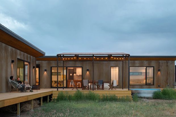 Single-storey contemporary residence viewed from a landscaped outdoor area, featuring a low horizontal form, extensive glazing, timber-clad walls, and a broad timber deck. A central covered outdoor dining/lounge zone has a slender pergola with warm string lighting, while the left and right wings frame the courtyard-like frontage. The setting is overcast and surrounded by natural grasses.